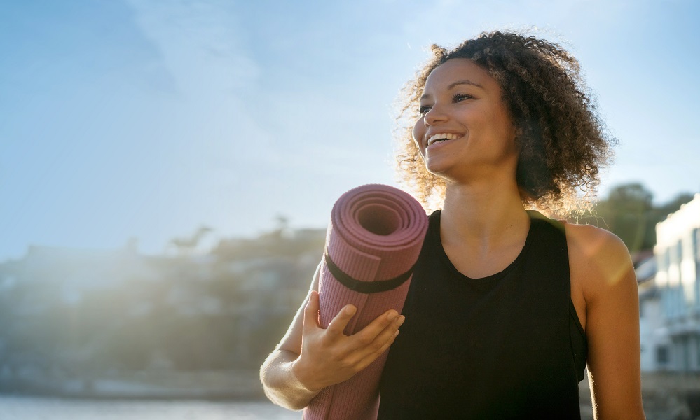 Portrait of a fit woman holding a yoga mat at the beach and looking very happy - wellness concepts