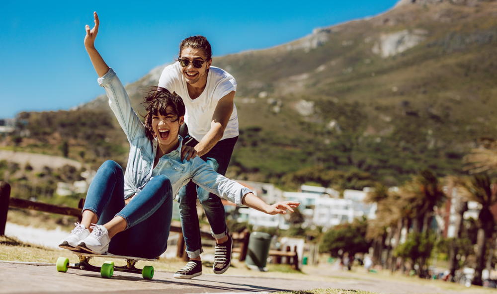 Couple having fun outdoors near the beach. Man pushing her girlfriend on a skateboard.